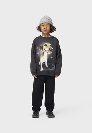 Kind trägt graue Mütze, schwarzen Pullover mit großem lachendem Hundemotiv, schwarze Hose und schwarze Schuhe, steht vor einem schlichten hellen Hintergrund.