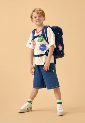 Shorts en jean bleu, T-shirt crème avec des imprimés de chats colorés, et un sac à dos bleu foncé avec des motifs spatiaux. Chaussures blanches avec des accents verts.