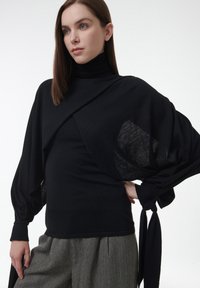 Schwarzes Langarmoberteil mit hohem Kragen, das über weite Ärmel, ein strukturiertes Panel und Bindebänder an den Handgelenken verfügt. Getragen mit gemusterten Hosen.