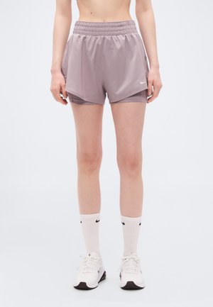 Άτομο που φοράει μοβ στρωμένες κοντές φόρμες τρεξίματος, λευκές κάλτσες Nike και λευκά αθλητικά παπούτσια, στέκεται μπροστά σε ένα απλό φωτεινό φόντο.