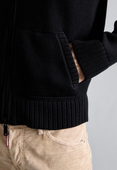 Zwarte ritssluitingjack met ribgebreide manchetten en zoom, gemaakt van een textuurgebreide stof en met voorzakken. Draag met beige corduroy broeken.