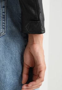 La manche d'une veste en cuir noir présente une finition texturée, avec un design de poignet pointu. Un tissu en denim bleu est visible en dessous de la manche.