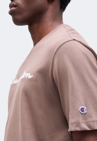 Vue de profil d'une personne portant un t-shirt marron Champion avec un logo brodé sur la manche et un texte partiel sur la poitrine.