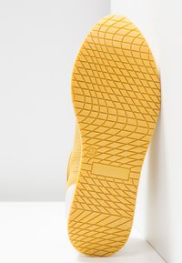 Semelle de chaussure en caoutchouc jaune avec un motif de bande de roulement géométrique, présentée debout contre un mur blanc d'angle.