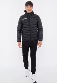 Svart puffjacka med horisontell sömnad, hel dragkedja, huva och vit logga på vänstra bröstet. Matchad med svarta byxor och vita sneakers.