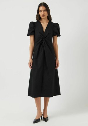 Vrouw staand, dragend een zwarte midi-jurk met korte pofmouwen, een gedraaide knoopdetail ter hoogte van de taille en zwarte puntige hakken.