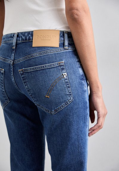 Pantalones vaqueros de mezclilla azul con una etiqueta de cuero de la marca, costuras detalladas en los bolsillos traseros y un corte relajado. La textura parece suave y durable.