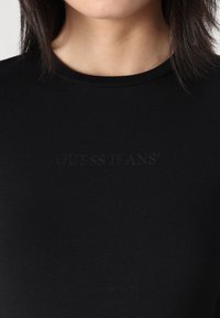 Černé přiléhavé tričko vyrobené z měkké tkaniny, s kulatým výstřihem a vystouplým logem "GUESS JEANS" na přední straně.