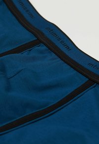 Modré boxerky s černým elastickým pasem, které mají hladkou texturu a logo značky podél horního okraje.