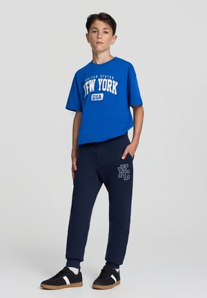 T-shirt blu a maniche corte con grafica "New York"; pantaloni della tuta blu scuro con logo; sneakers nere con strisce bianche; vestibilità casual.