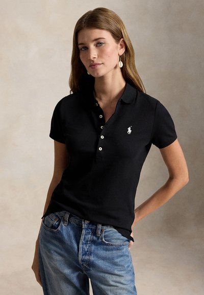 Kvinna med långt hår iförd svart pikétröja med vit hästlogotyp och blå jeans, står med ena handen i fickan.