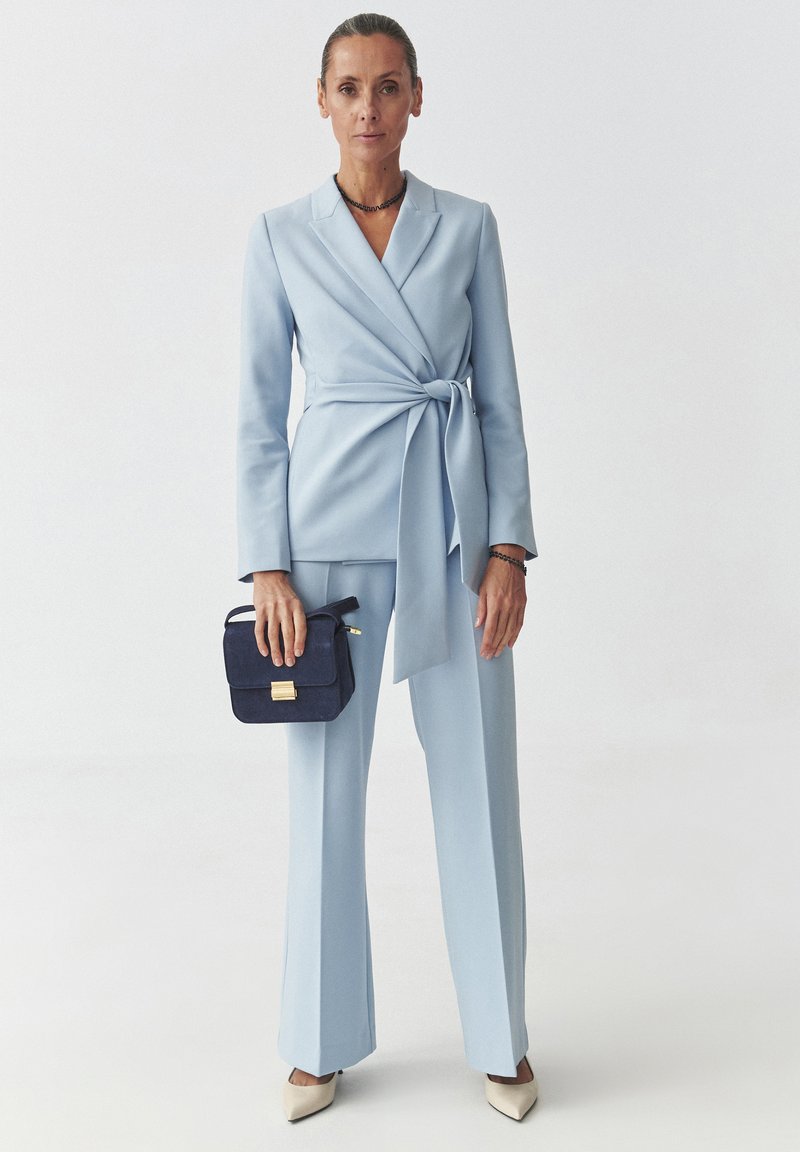 Helles blaues Tailoring-Anzug mit Wickeldesign, am Taillieren gebunden. Beigefarbene Absatzschuhe und eine kleine marineblaue Umhängetasche mit goldenen Details.