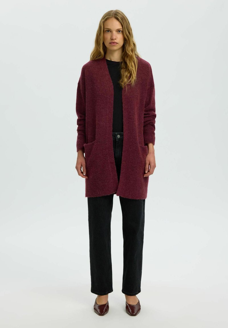 Femme debout portant un long cardigan bordeaux avec poches avant, un haut noir, un pantalon noir et des chaussures bordeaux à bouts pointus sur un fond uni.