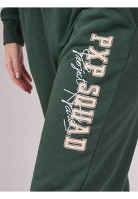 Groene trainingsbroeken hebben de tekst "PXP SQUAD" in beige en wit aan de zijkant, met een zachte stof en een comfortabele pasvorm.