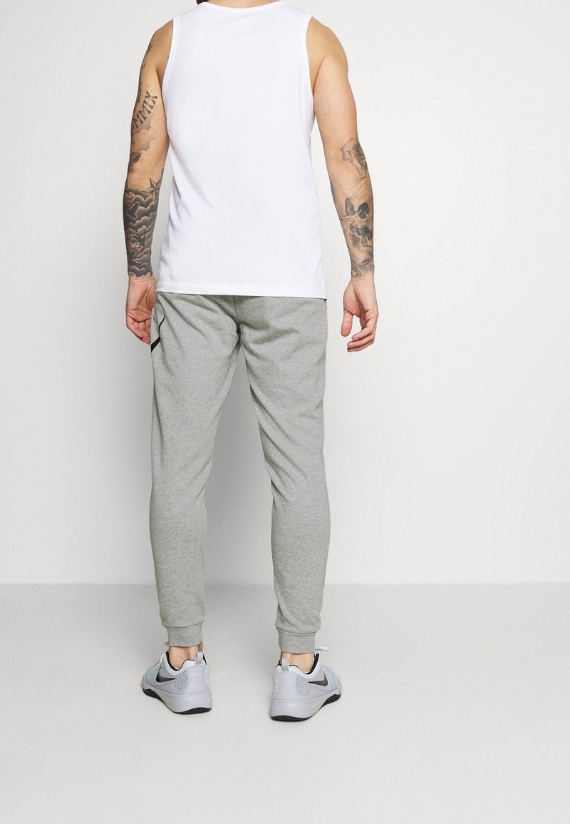 Homme aux bras tatoués portant un débardeur blanc, un pantalon de survêtement gris et des chaussures de sport grises, debout de dos.