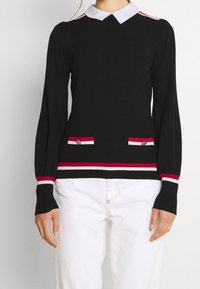 Schwarzer Strickpullover mit weißem Kragen, rot-weiß gestreiften Akzenten an Ärmeln und Saum, mit zwei vorderen Taschen und goldenen Knöpfen.