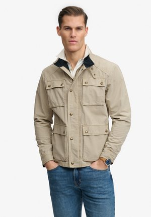 Mand iført beige jakke med flere klaplommer, hvid skjorte med krave, blå jeans og armbåndsur, stående med hænderne i lommerne.