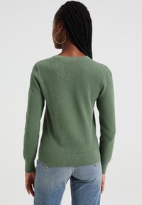 Green textured sweater with long sleeves and a round neckline. Features a ribbed hem and cuffs, paired with light blue jeans.