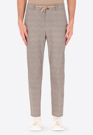 Uomo che indossa pantaloni beige con coulisse e trama, e sneakers bianche, in piedi con le mani rilassate ai lati.