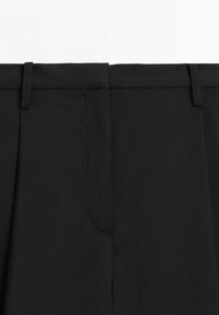 Detailní záběr oblasti pasu černých kalhot s poutky na pás a skrytým předním zapínáním s pečlivými šicími detaily.