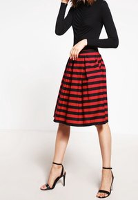 Femme portant un haut noir à manches longues, une jupe rouge et noire à rayures horizontales arrivant aux genoux, et des sandales à talons hauts noires.