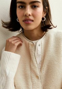 Krämfärgad, texturerad fleeceväst med ribbstickade ärmar, knäpplås och en mjuk inre foder. Detaljer inkluderar en rund halsringning.