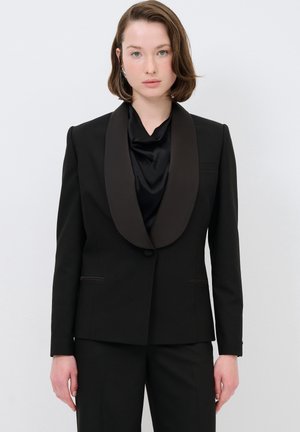 Schwarzer maßgeschneiderter Blazer mit einem satinbachenden Revers, ausgestattet mit einem einzigen Knopfverschluss und zwei Vordertaschen, getragen über einem schwarzen Satinoberteil.