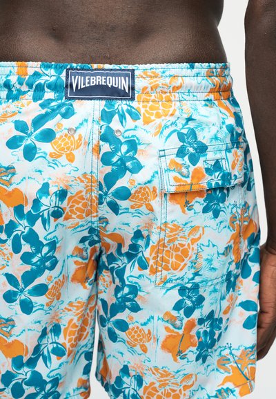Μαγιό σορτς με πορτοκαλί και τιρκουάζ floral και σχέδιο με χελώνες, πίσω τσέπη με καπάκι και ετικέτα "Vilebrequin" στη μέση, φορεμένο από άτομο.