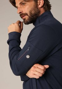 Marine blauwe zip-up jas met een hoge kraag, voorzien van een ritssluiting op de linkerarm en een metalen accentknop. Glad oppervlak.