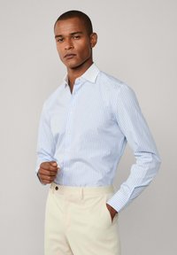 Uomo che indossa una camicia a righe blu chiaro e bianca con colletto bianco e pantaloni avorio, in piedi contro un muro semplice.