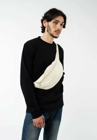 Beige Stoff Umhängetasche mit kurvenförmiger Silhouette, vorderer Reißverschluss-Tasche, verstellbarem Tragegurt und geprägtem Logo, quer über dem Körper getragen.