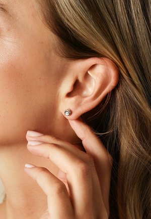 Nahaufnahme einer Hand, die ein Ohr mit einem silbernen Steckerohrring mit einem klaren Edelstein berührt, wobei Strähnen von hellbraunem Haar sichtbar sind.