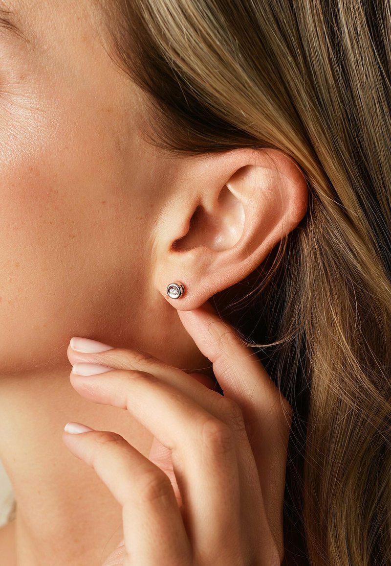 Nahaufnahme einer Hand, die ein Ohr mit einem silbernen Steckerohrring mit einem klaren Edelstein berührt, wobei Strähnen von hellbraunem Haar sichtbar sind.