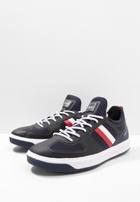 Tummansinisiä urheilukenkiä, joissa on valkoiset ja punaiset raidat, verkkomaisia paneeleja, valkoiset nauhat ja paksu valkoinen pohja, sekä logo kantapäässä.