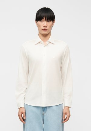 Mand med sort hår iført en hvid langærmet skjorte med knapper og lyseblå jeans foran en ensfarvet baggrund.