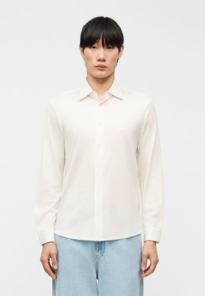 Hombre con cabello negro que lleva una camisa blanca de manga larga abotonada y jeans azul claro, sobre un fondo liso.