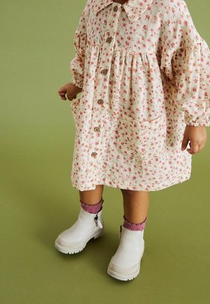 Robe crème à motif floral avec manches bouffantes, boutonnée à l'avant et poches latérales. Bottines blanches à fermeture éclair latérale et chaussettes roses.