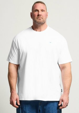 Muskelbepackter Mann mit kurzen Haaren und Bart steht frontwärts, trägt ein schlichtes weißes T-Shirt und blaue Jeans vor hellem Hintergrund.