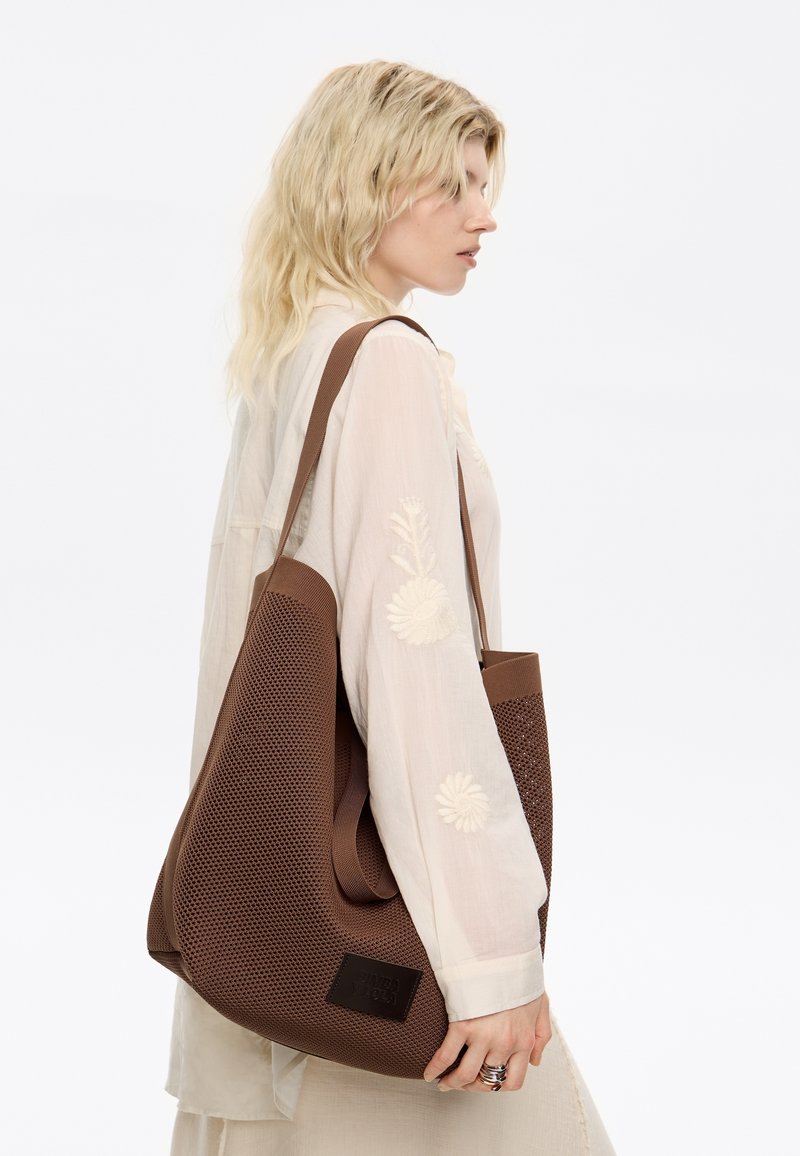 Borsa tote perforata marrone con manici larghi, dotata di una patch nera rettangolare con logo. Il modello mostra la borsa in un angolo laterale.