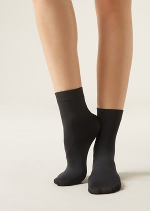 Nohy v jednoduchých černých kotníkových ponožkách stojící na světlém neutrálním pozadí, jedna noha mírně zvednutá na špičkách.