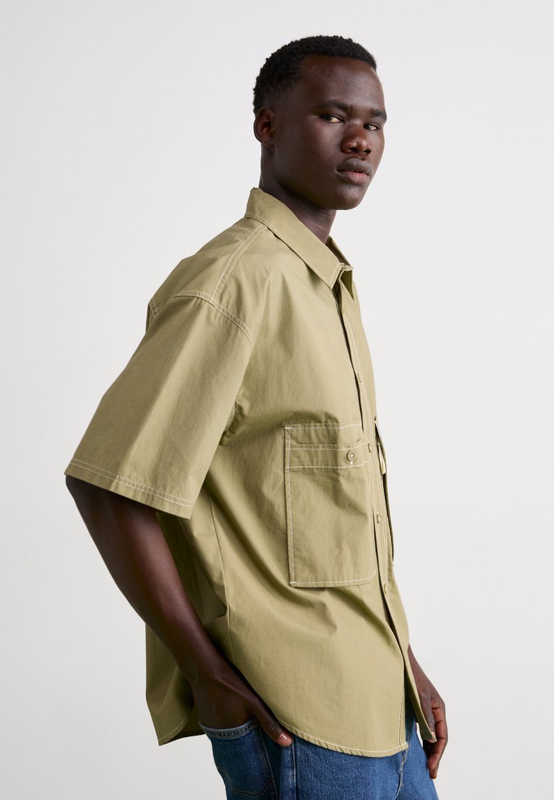 Camisa de manga corta en algodón verde oliva claro con cuello puntiagudo, bolsillos en las costuras laterales y detalles de costura en contraste.