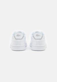 Valkoiset nahkakengät, joissa on sileä pinta, pehmustettu kaulus, pyöreä kärki ja hopea Lacoste-logo kantapäässä. Näkyvillä oleva kumipohja.