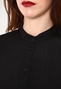 Mustassa kauluspaita, jonka kankaassa on tekstuuria. Paidassa on napitettu etuosa ja kevyt kiilto, mikä korostaa sen siistiä ja minimalistista muotoilua.