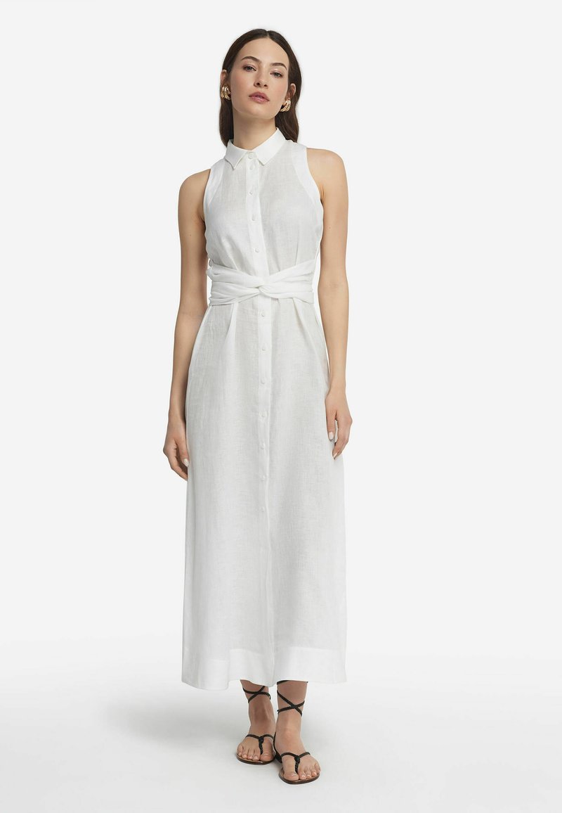 Weißes Leinenhemdkleid mit Kragen und Knopfleiste vorne. Hat eine geknotete Schleife in der Taille. Modell trägt schwarze Riemchensandalen.