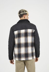 Podložena črna jakna iz tkanine s karirasto hrbtno stranjo v odtenkih črne, sivo-bež in smetane. Ima ovratnik in teksturirane detajle.