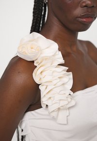 Weißes Kleid mit einem gerafften Stoffakzent an der Schulter, hergestellt aus glattem Material mit einem sanften Glanz. Nahaufnahme von Textur und Detail.