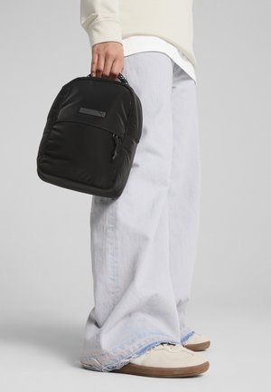 Zwarte gewatteerde rugzak met een voorzak met rits en een gestructureerde band, vastgehouden door een persoon in lichtgekleurde wijde jeans en beige sneakers.