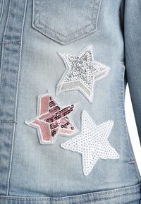 Kurtka jeansowa z trzema naszywkami w kształcie gwiazd: jedna ze srebrnych cekinów, druga różowa z cekinami, a trzecia biała z koralikami, wszystkie z białymi obwódkami.