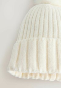 Kreminė trijų modelių megzta skrybėlė su vertikaliais raukiniais, minkšta tekstūra ir apverstu kraštu. Viršuje yra pūkuotas pom-pom.