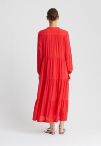 Robe rouge à manches longues avec une silhouette fluide à volants. Confectionnée dans un tissu léger, présentant un dos froncé et un col rond. Sandales visibles.